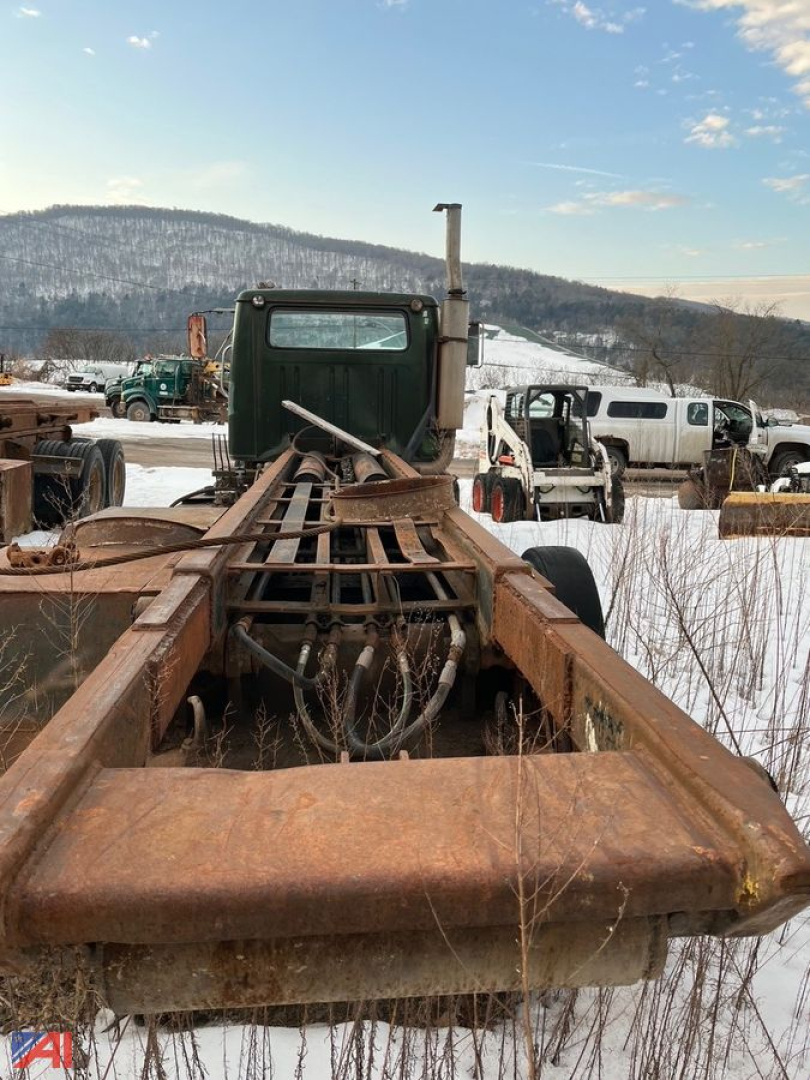 Truck chassis and cab of an International roll-off truck with rusted frame, winter surroundings, and a tall exhaust stack behind the cab.