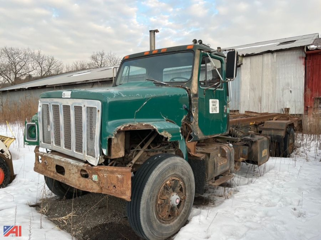 1986 International 2554 Roll Off Truck with green cab and exposed rusted front bumper, side view in snow, worn body panels and missing bed.