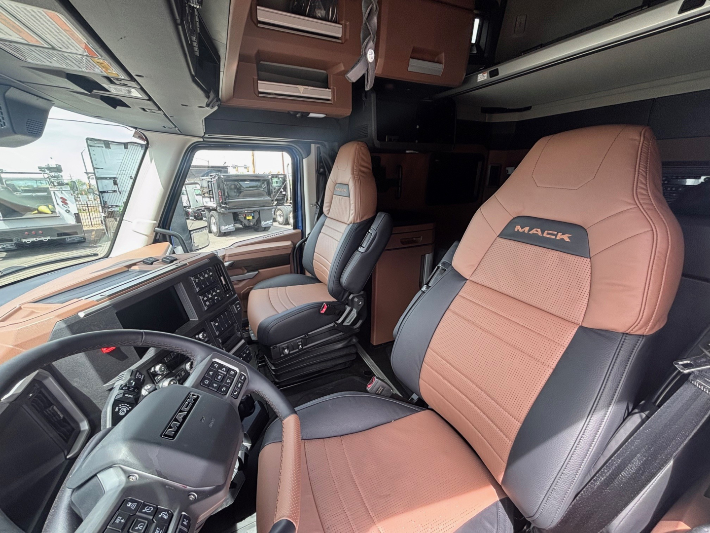 Truck cockpit interior of a 2027 Mack PR64T76H showing tan and black two-tone seats with Mack logo, steering wheel, dashboard, and sleeper cab area.