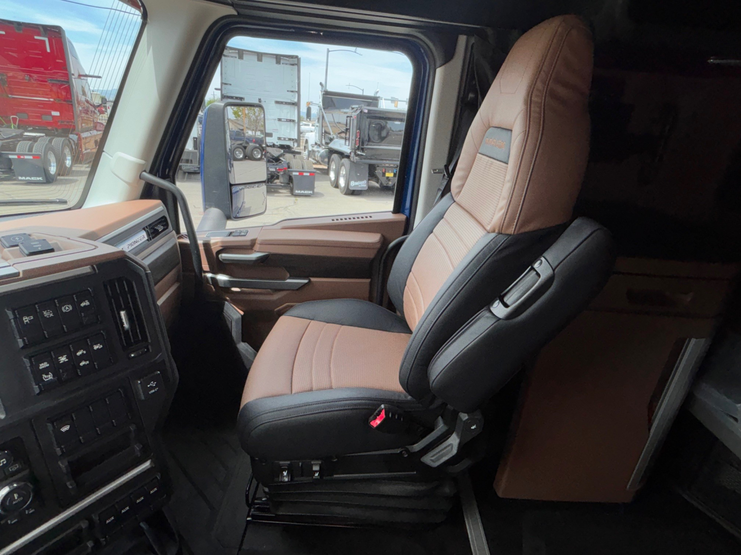 Cab interior of a 2027 Mack PR64T76H. Front driver seat with tan and black upholstery, dashboard and door panel visible, exterior trucks seen through window.