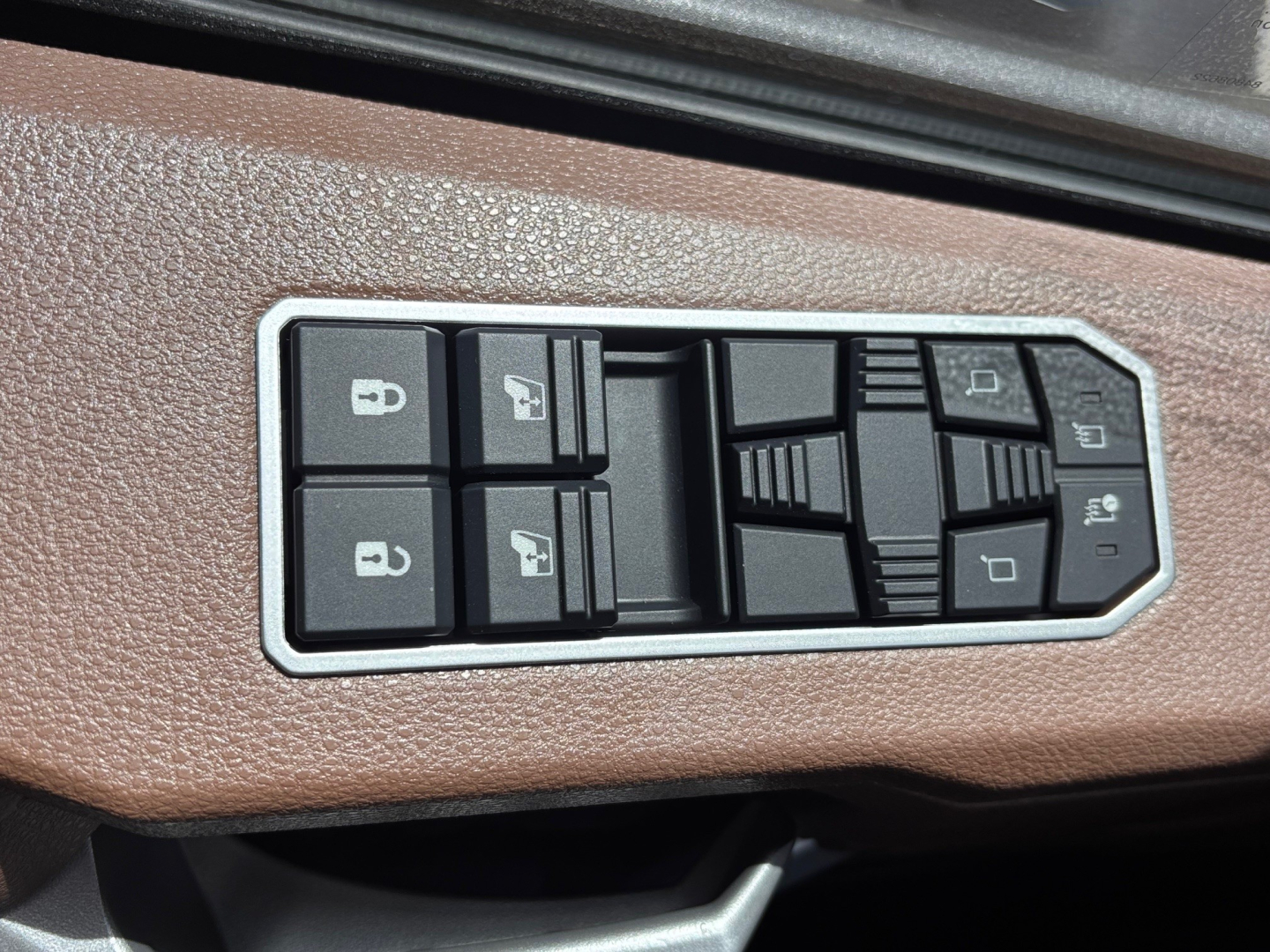 Inside cab window/door panel with multiple control switches: four square buttons with window icons and two rows of smaller gray rocker switches on brown textured panel.