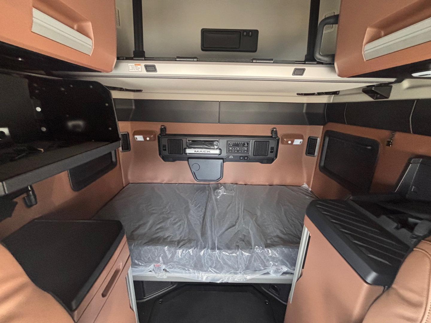 Truck cab interior showing sleeper area with overhead storage, control panel, and brown leather-like upholstery; center control dash; bed area covered with protective plastic.