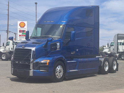 Truck cab showing a blue Mack PR64T76H tractor at a dealership lot with sleeper cab and chrome wheels.
