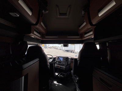 Truck cab interior of a 2027 Mack PR64T76H viewed from back toward the dashboard, seating and overhead storage visible.