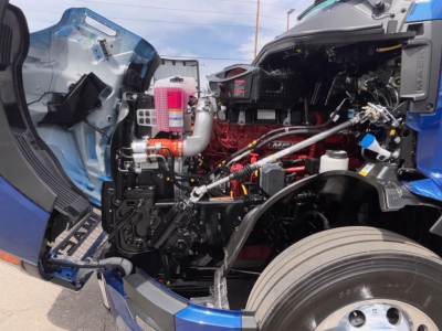 Mack PR64T76H engine bay showing red engine block, intake piping, pink coolant reservoir, black chassis, cab interior visible, front wheel and fender.