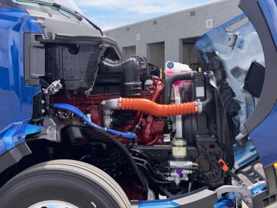Truck engine bay of a 2027 Mack PR64T76H with blue cab, showing orange coolant hose, red engine, blue hoses, and various components.