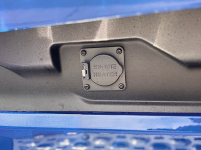 Truck engine bay access panel labeled ENGINE HEATER on the cover in a blue Mack PR64T76H.