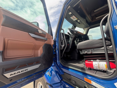 Mack PR64T76H truck cab interior showing driver seat and open door with 'MACK' branding on door sill and a fire extinguisher nearby.
