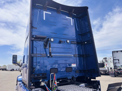 Truck cab rear view of a blue 2027 Mack PR64T76H conventional sleeper showing the back of the sleeper, tailgate hinges, frame, and wiring harnesses.