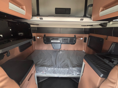 Truck cab interior showing sleeper area with overhead storage, control panel, and brown leather-like upholstery; center control dash; bed area covered with protective plastic.