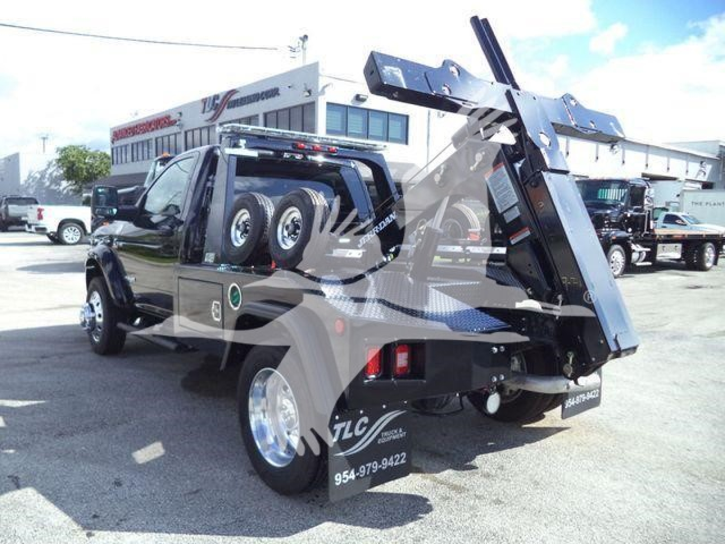 Truck with a heavy-duty tow/roll-off hardware system mounted in the bed, dual rear wheels, and a hydraulic tilting crane/ramp. Visible text on rear includes TLC and contact number on fender; background shows a dealership lot.