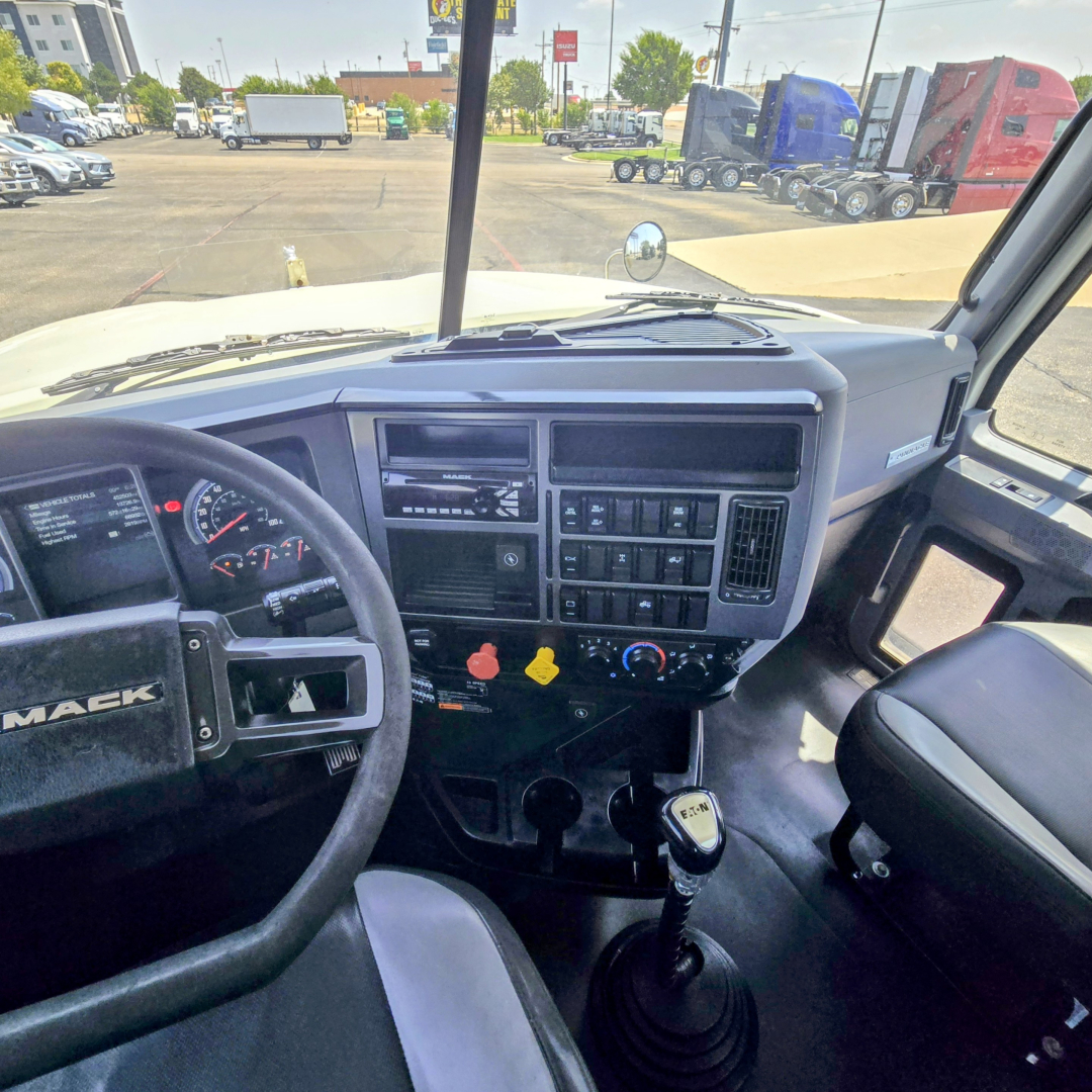 Mack PI64T cab interior showing steering wheel and dashboard controls with manual shifter and center console, view from passenger seat.