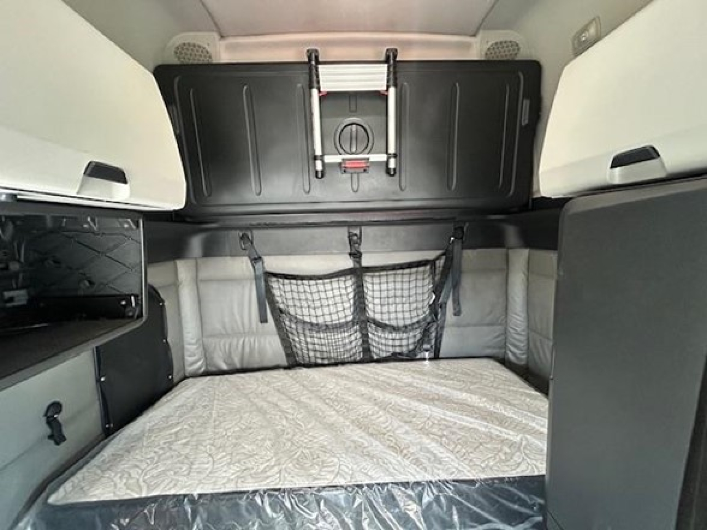 Freightliner Cascadia interior sleeper area showing bunk storage under cabinet and netted pocket, light gray upholstery and overhead panels.