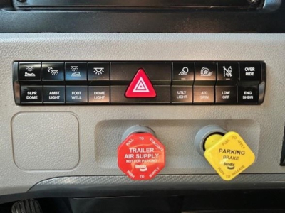 Dashboard controls of a Freightliner Cascadia 126 showing hazard switch, various indicator buttons, red trailer air supply knob, and yellow parking brake knob.