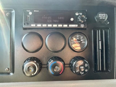 Dashboard panel of a Freightliner Cascadia showing a mono radio display, three circular gauges, climate controls, and a power outlet.