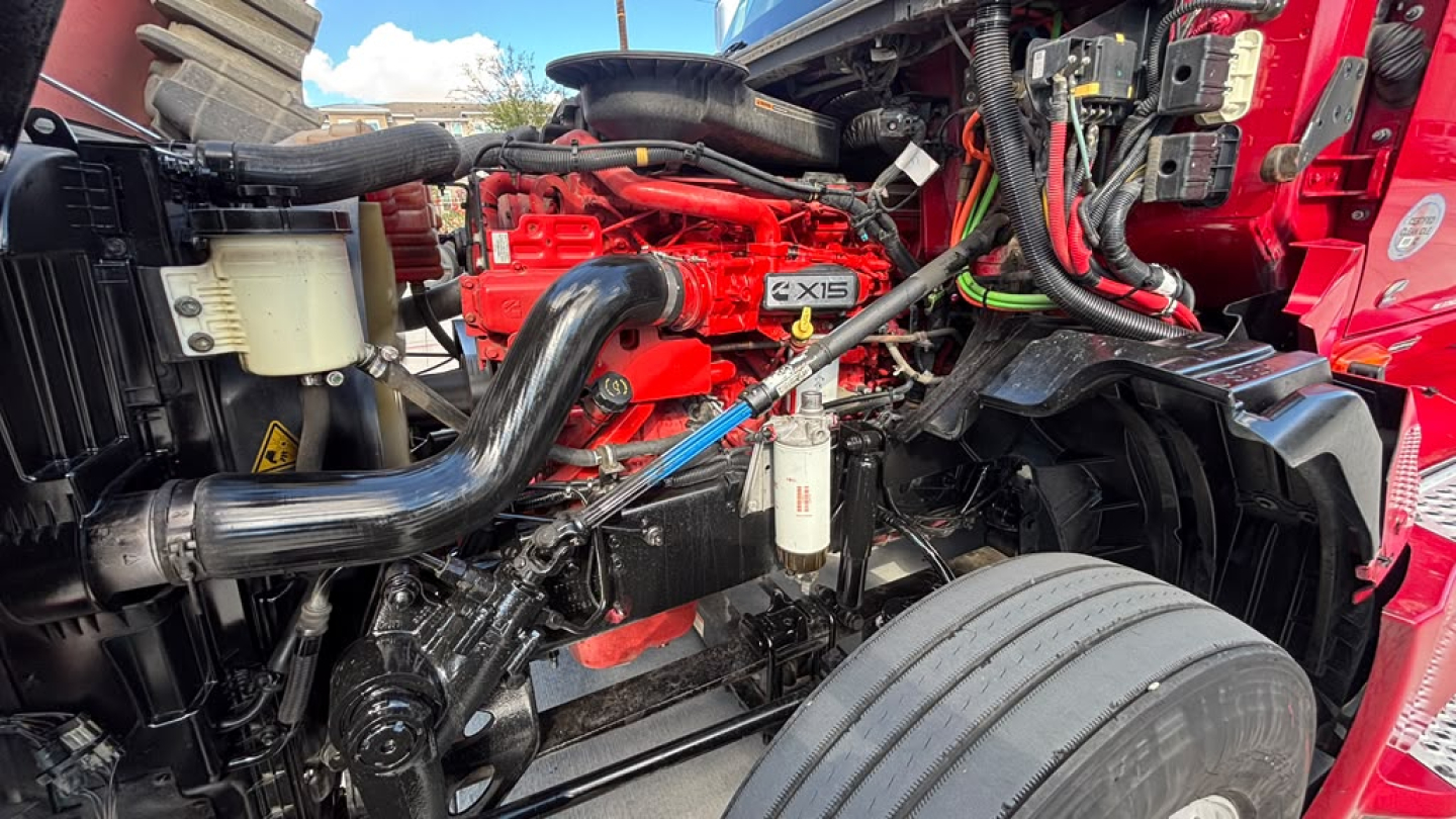 Truck engine bay showing International LT625 diesel engine with red-painted components, black hoses, and a large front wheel area; visible components include intake tubing, filters, and wiring harnesses.