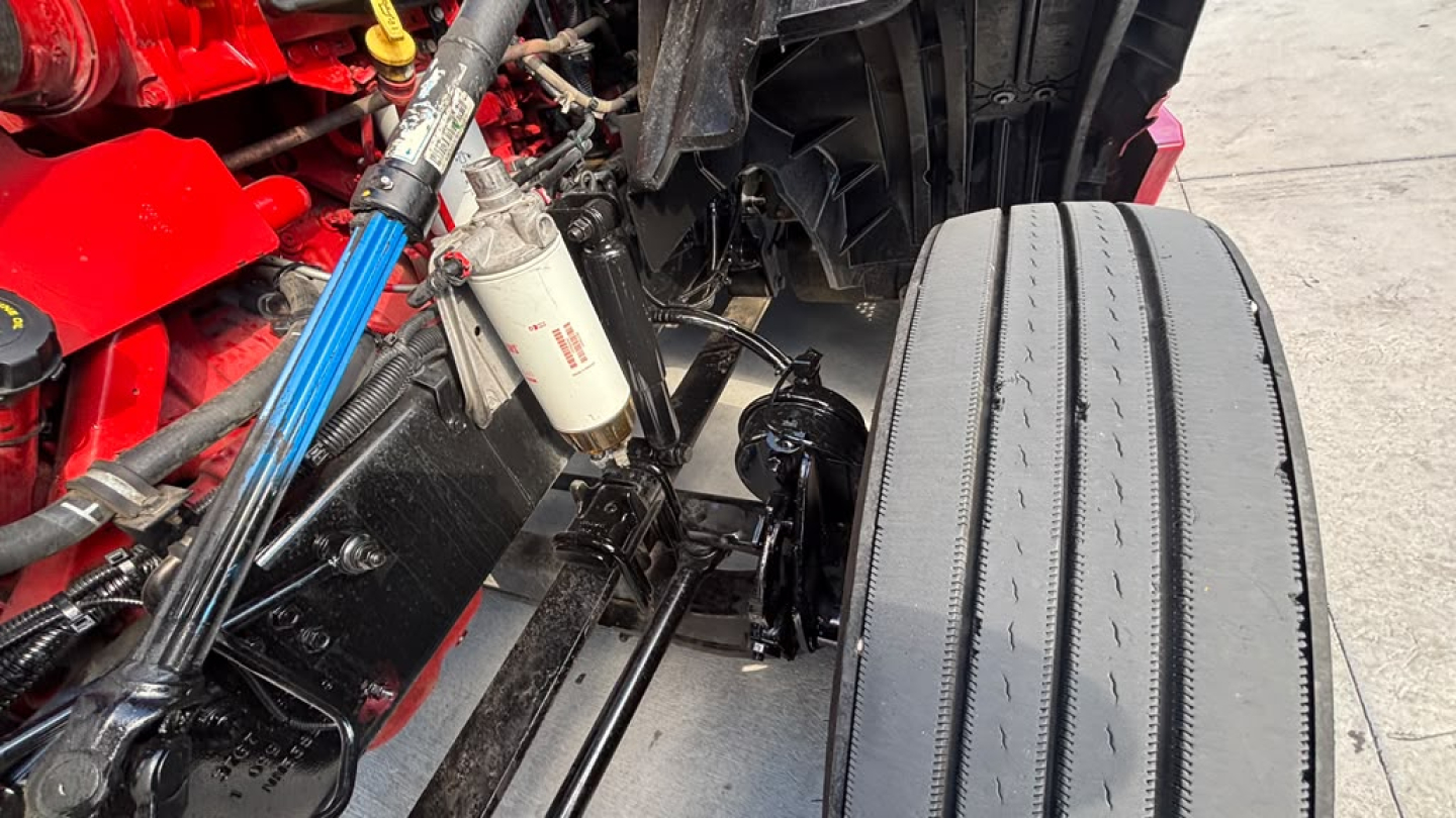 Truck engine bay view showing a red engine, white oil filter, blue hose, and a black suspension/axle area near the front tire.