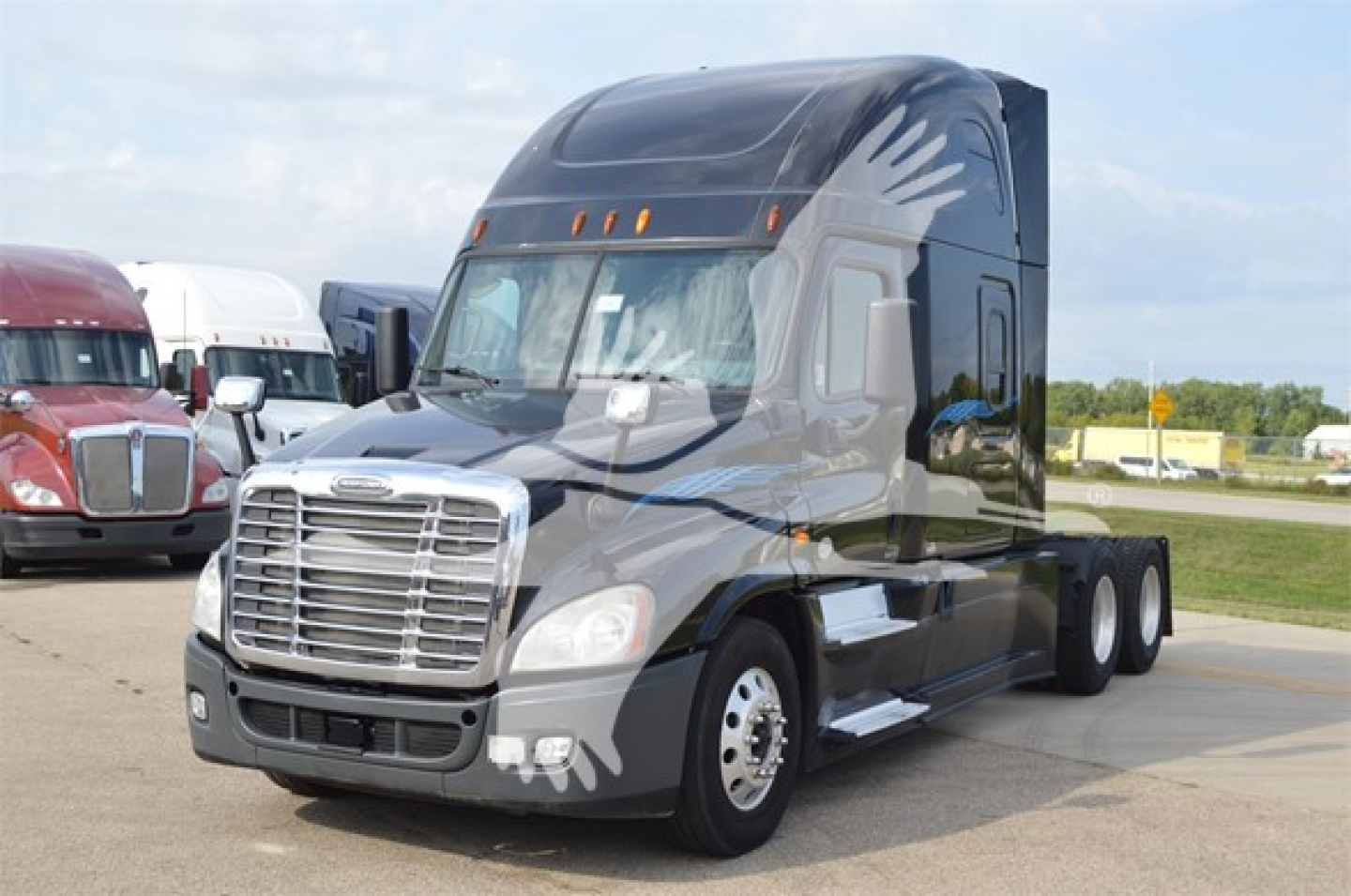 Truck cab with Freightliner emblem showing front grille and headlights; dark gray paint with aerodynamic sleeper cab in a lot.