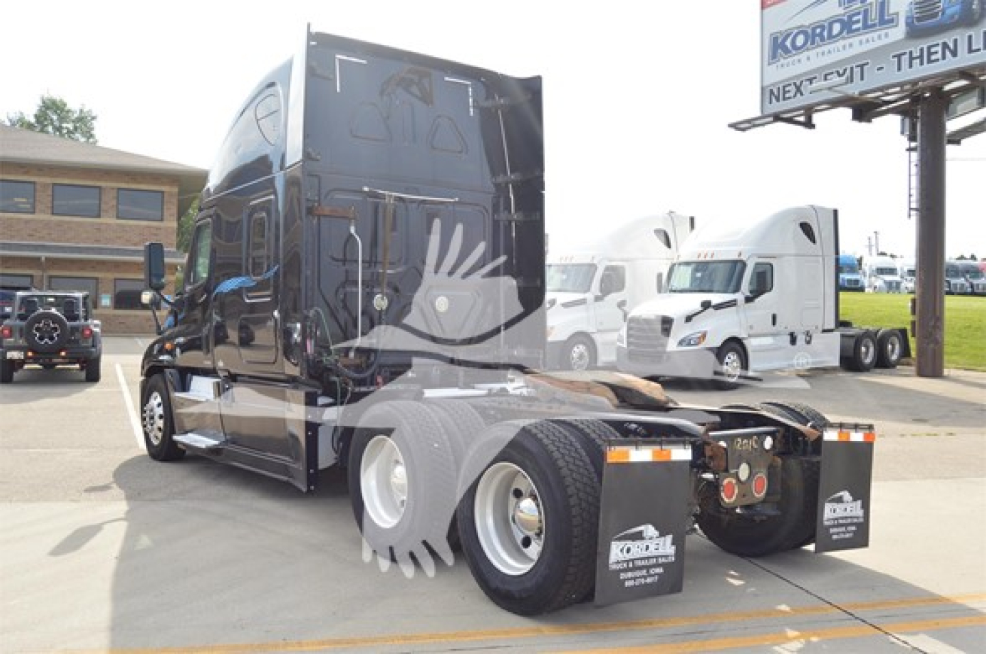 Truck rear view showing fifth wheel and dual rear axles, Freightliner badging not visible, reflective surface, parking lot with other trucks.