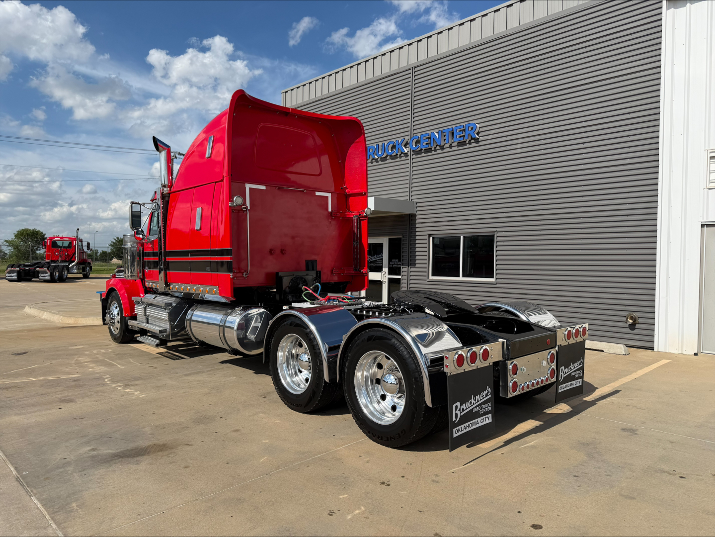 2020 Western Star 4900 conventional sleeper truck from rear-left side showing red cab, chrome fuel tanks, dual rear axles, and trailer hitch, dealership building in background.