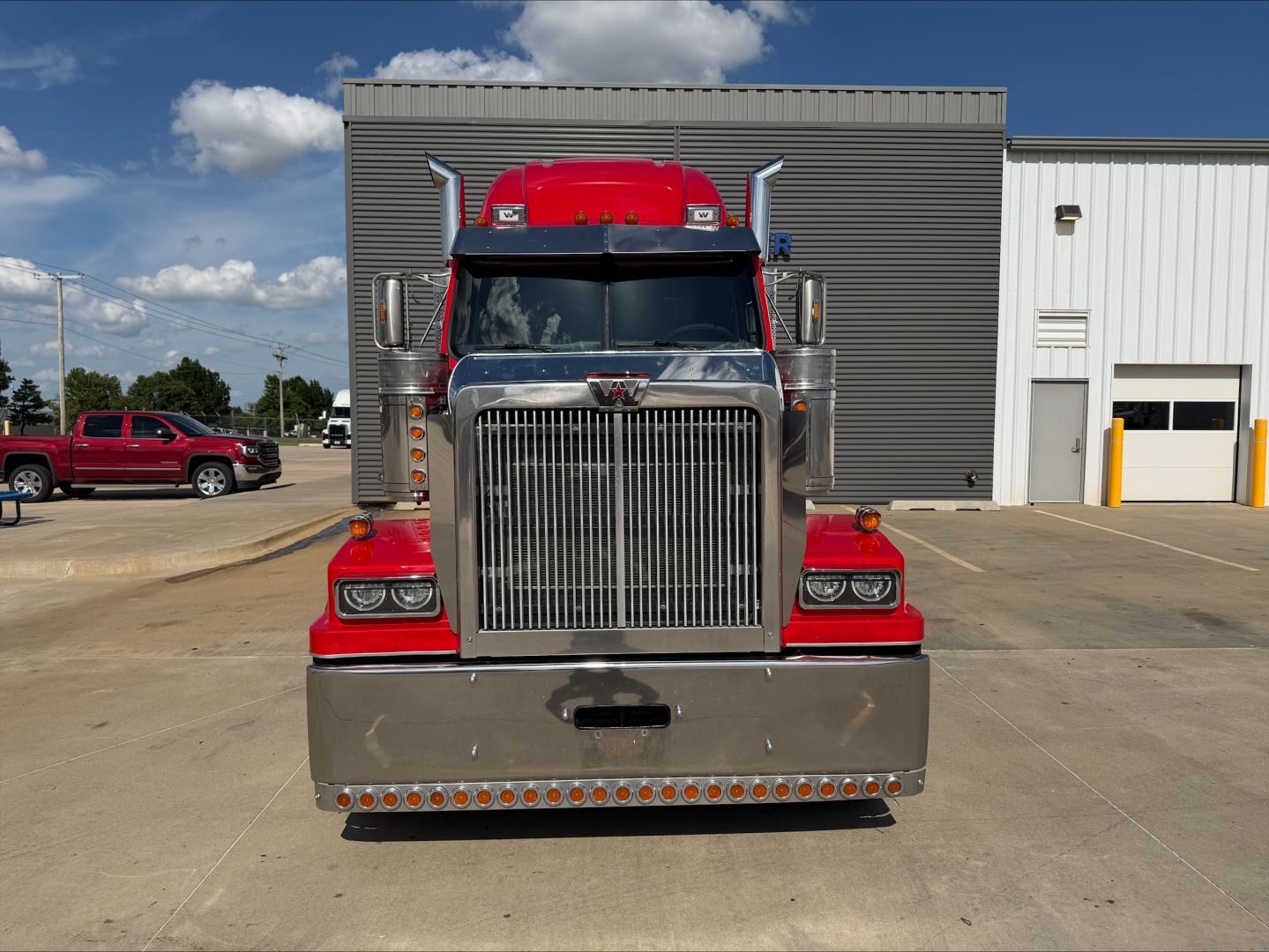 Front view of a red and chrome Western Star 4900 truck, large vertical grille, bumper with many small lights, two stacked headlights, tall exhaust stacks, on a concrete lot.