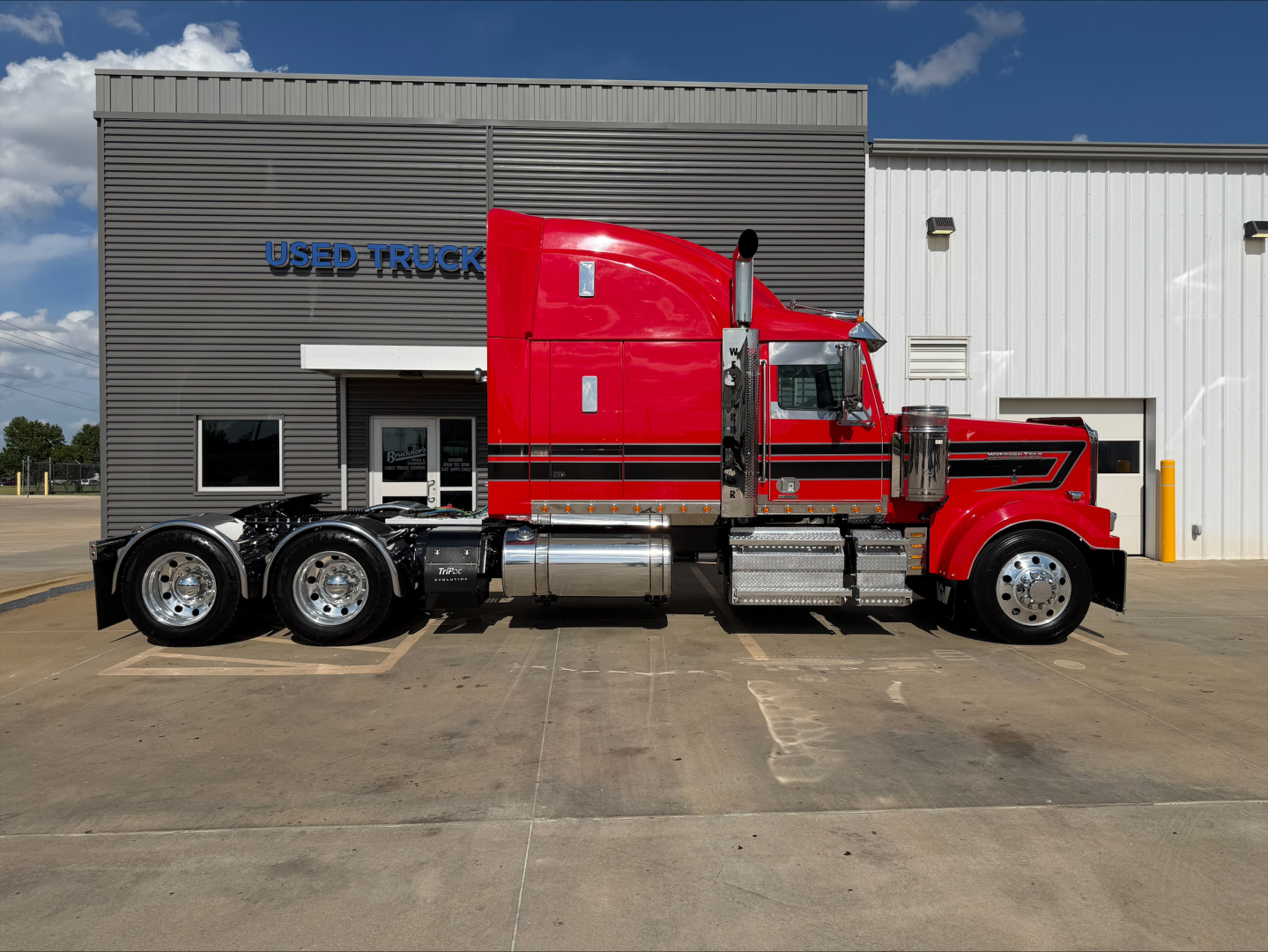Side view of a red 2020 Western Star 4900 conventional sleeper truck with chrome fuel tanks and dual rear axles, parked in front of a used trucks building.
