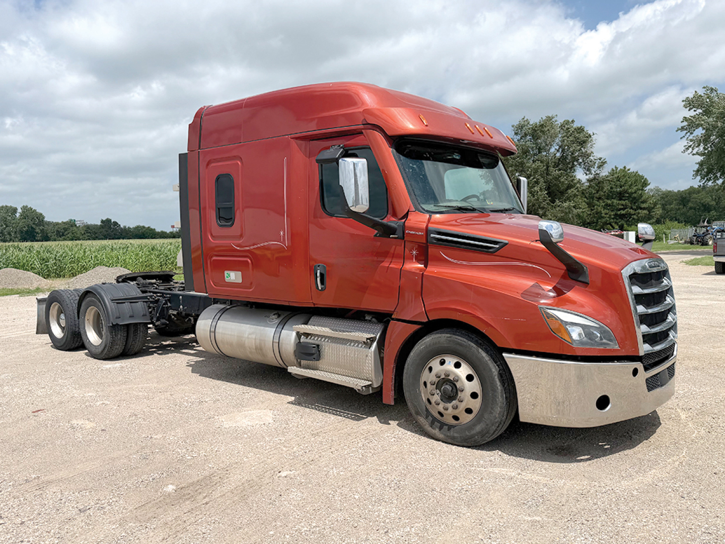Red Freightliner Cascadia126 cab truck in a gravel lot, side view showing fuel tanks, front bumper, and wheels.