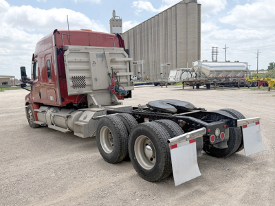 Freightliner Cascadia semi-truck tractor rear view showing fifth wheel, dual rear axles, and mud flaps in a gravel lot.