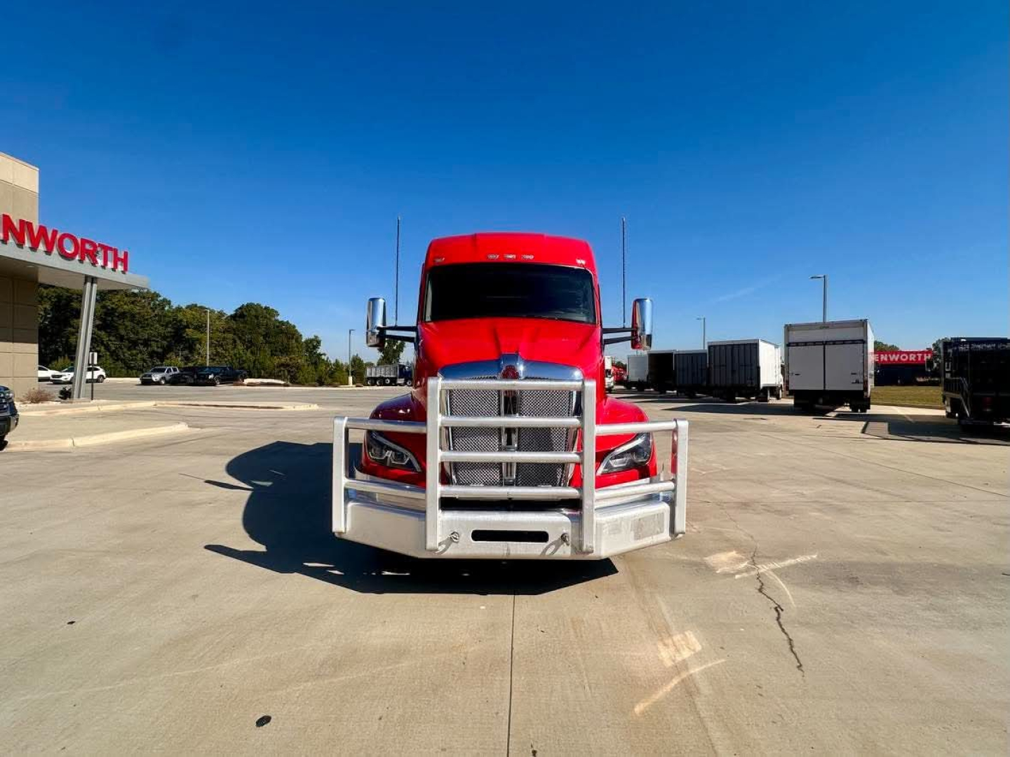 Kenworth T680 front view showing bold grille, bumper guard, and red cab with chrome bumper in a truck lot.
