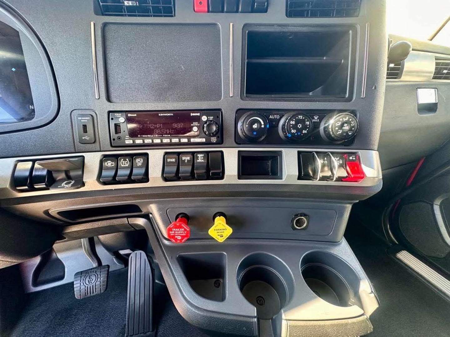 Kenworth T680 interior dashboard view showing center stack with radio, climate controls, red and yellow hazard knobs, and cup holders.