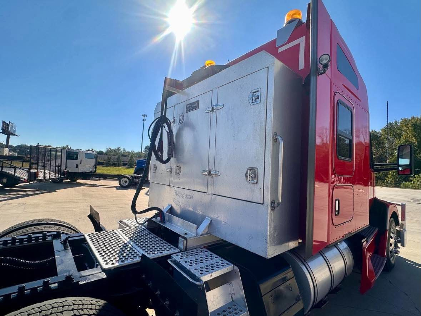 Kenworth T680 sleeper cab rear side showing stainless toolbox and exhaust area with fuel tanks, red cab, and roadside lot.