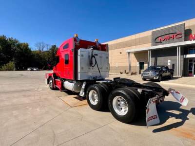 Kenworth T680 dual rear axles chassis in red color with sleeper cab, shown from rear-left side in parking lot; vehicle features and dealership building in background.