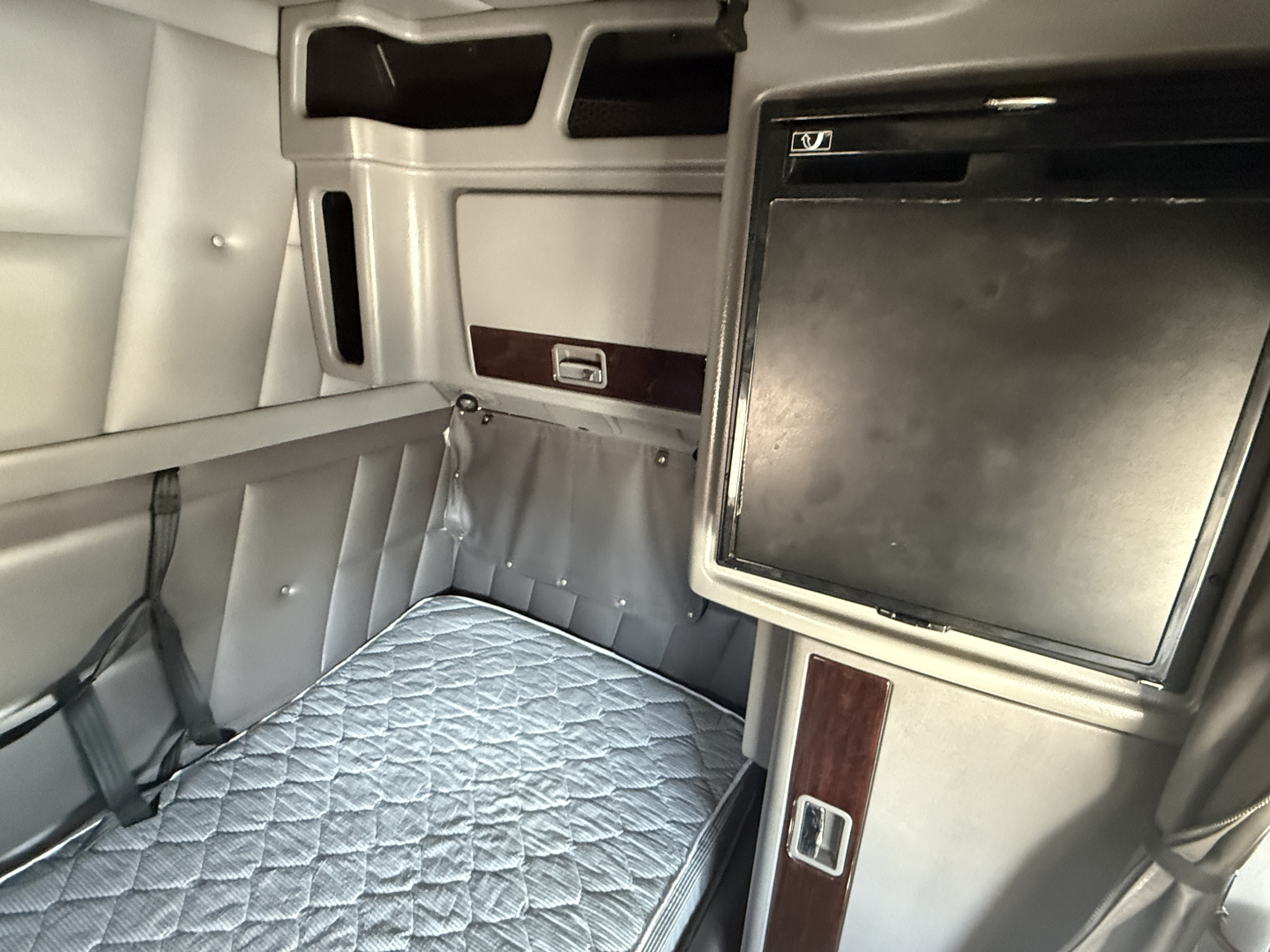Peterbilt sleeper cab interior showing bunk bed with quilted mattress, overhead cabinets, and a small fridge/locker with wood trim.