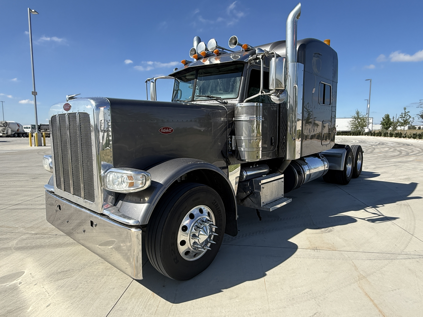 Peterbilt 389 conventional sleeper truck in dark color with chrome accents, showing front grille, large chrome bumper, dual exhaust stacks, and dual rear axles.