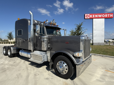 Peterbilt 389 conventional sleeper truck in front of Kenworth sign. Chrome exhaust stacks, fuel tanks, and aluminum steps visible. No. plate and dealership text not legible.
