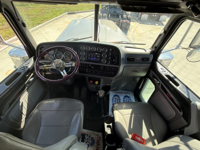 Truck cab interior of a 2020 Peterbilt 389 viewed from the driver door, showing steering wheel, gauge cluster, dashboard, gear shifter, and two seats.