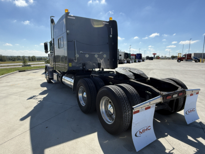 Peterbilt 389 tractor rear view showing dual rear axles and mud flaps with MHC logo, blue sky, highway trucks in background.
