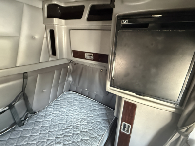 Peterbilt sleeper cab interior showing bunk bed with quilted mattress, overhead cabinets, and a small fridge/locker with wood trim.