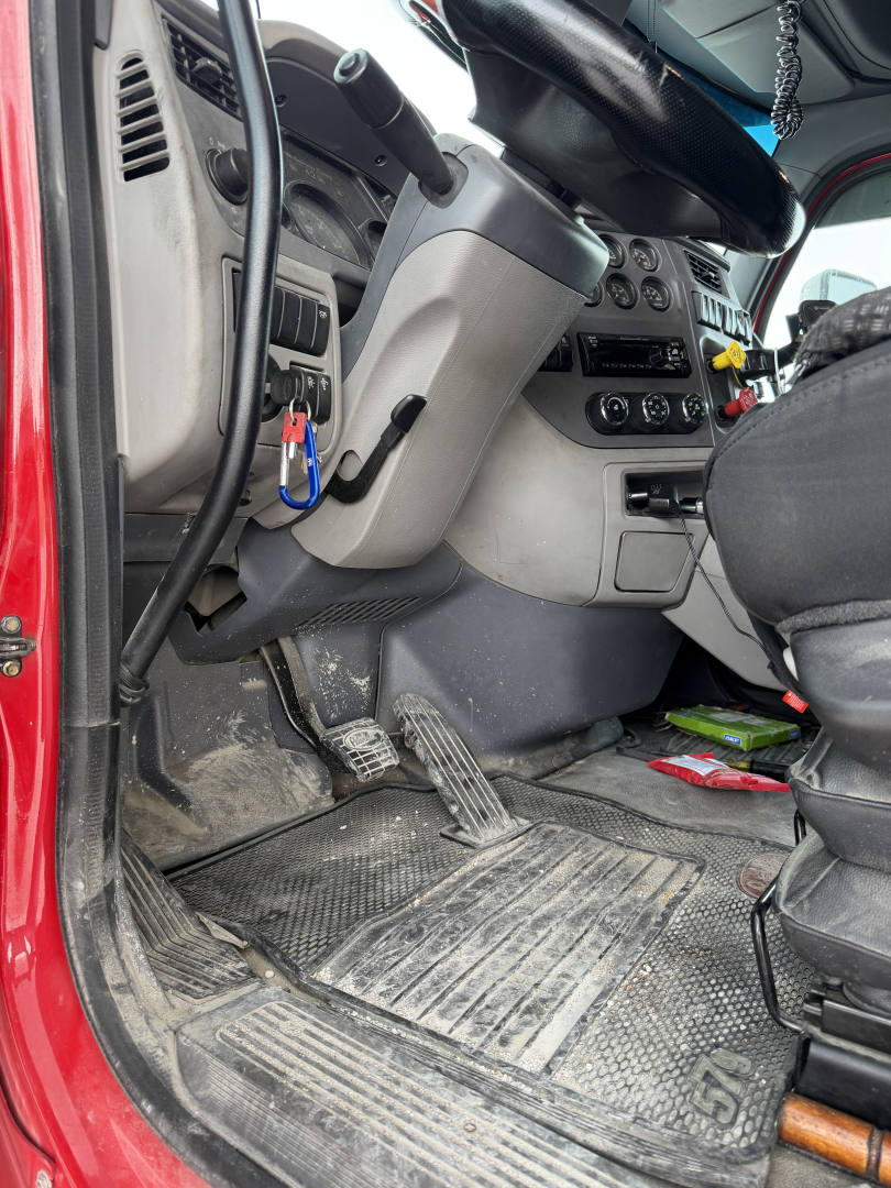 Truck cab interior of a 2017 Peterbilt 579 showing driver seat, dashboard, steering column, and floor area with pedals and exposed wiring.