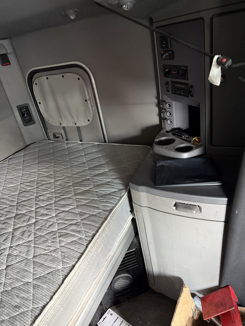 Peterbilt 579 sleeper cab interior showing bed with quilted mattress, controls panel, cup holder, and storage cabinet.