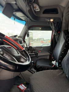 Truck interior showing steering wheel, dashboard, driver's seat, and cabin area of a Peterbilt 579 cab with accessories and hanging items.