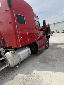 Side view of a red Peterbilt 579 sleeper cab truck showing fuel tank, door with text, and window; visible text includes 'NEWSOM TRUCKING INC' and other small lettering near cab door.