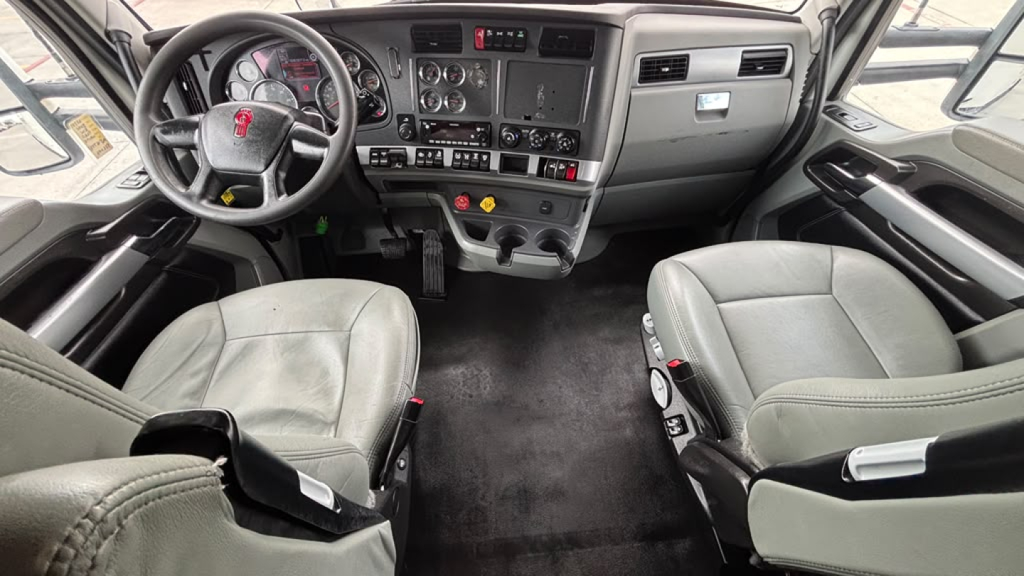 Kenworth T680 cab interior showing steering wheel, dashboard with gauges, center console, two seats, and floor mats.