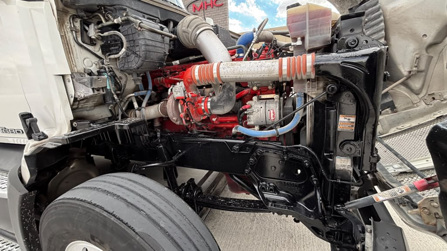 Kenworth T680 engine bay showing diesel engine with red components and black frame, front wheel, hoses, and radiator area.