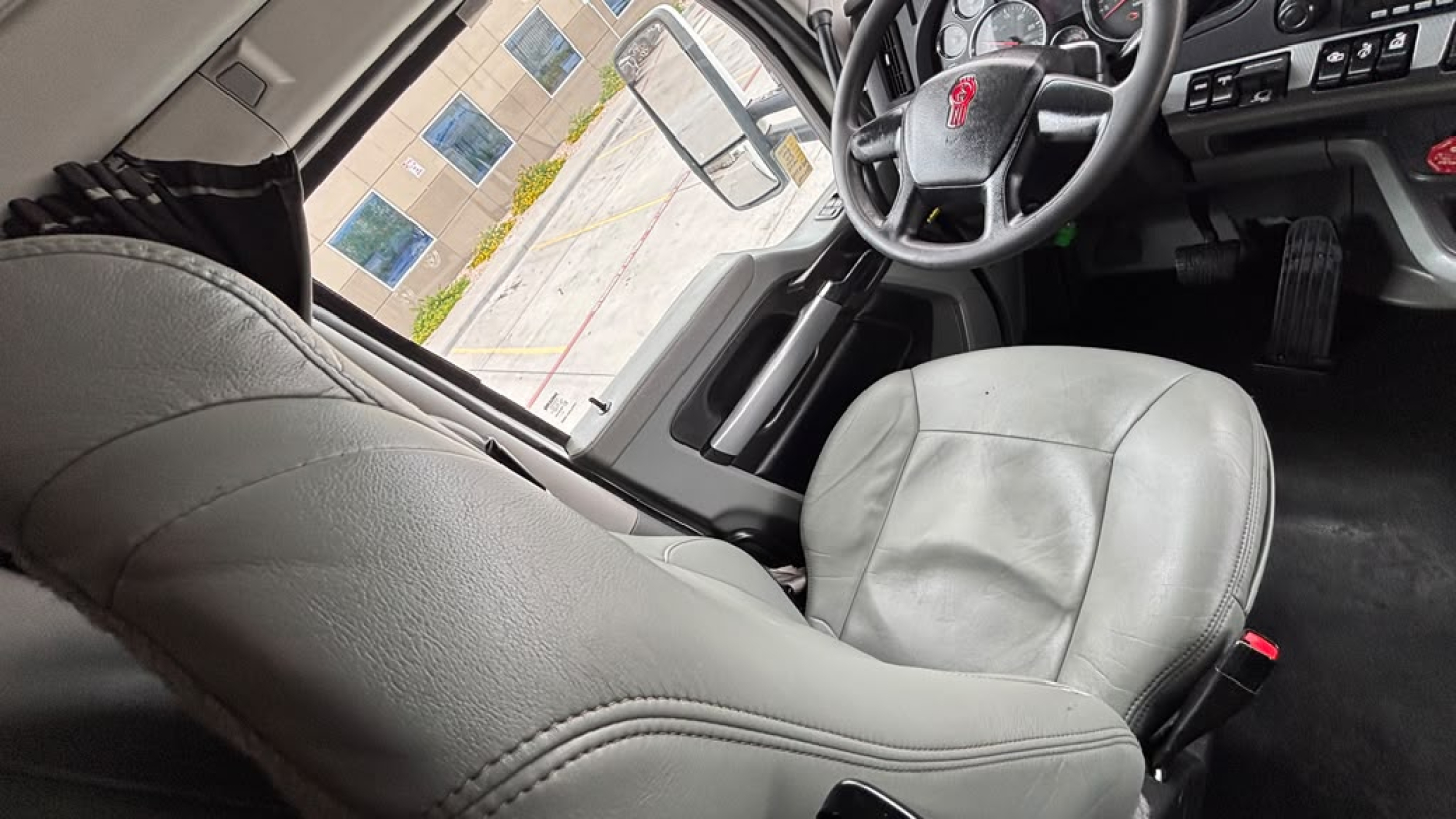 Kenworth T680 cab interior front seats and steering wheel, light gray upholstery, dashboard and pedals visible, driver seat close-up