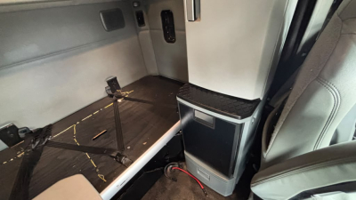 Kenworth T680 cab interior showing sleeper area with wood-look floor, small cabinet, and swivel seat; visible wiring and panel edges.