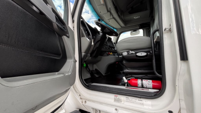 Kenworth T680 cab interior viewed from open driver door showing steering wheel, dashboard, and red fire extinguisher in footwell.