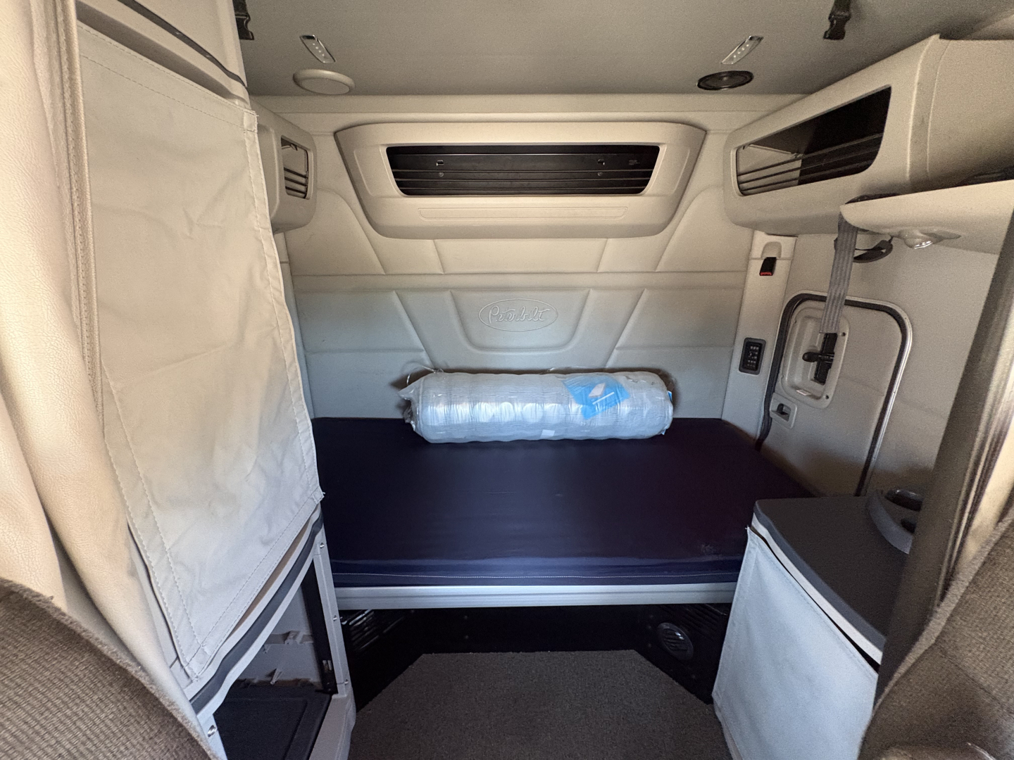Peterbilt 579 truck cabin interior shown in sleeper area with white sides, overhead cabinets, and a rolled mattress in the back, cargo space visible.