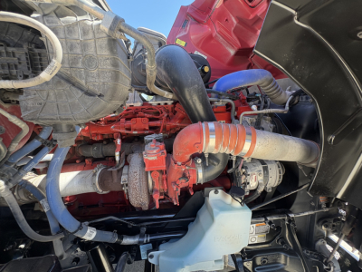 Engine bay of a Peterbilt truck with red engine, blue hoses, and black fender. Visible components include a large air intake hose, coolant reservoir cap, and various hoses and metal fittings.