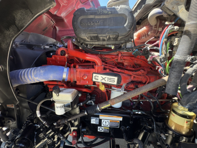 Peterbilt 579 engine bay showing Cummins X15 engine with red paint, blue radiator hose, white brake reservoir and various hoses and wiring in view.